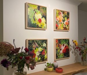 A series of abstract botanical paintings hang on a wall. Each painting is framed in a natural wood floater frame, and the paintings are arranged in a square above the mantelpiece. The dominant colours are red, green and yellow.